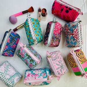 以下が含まれることがあります： 花柄や模様が施された、カラフルなキルティングコスメポーチのコレクション。ポーチにはファスナーと小さなハンドルが付いています。メイクブラシやアプリケーターも写っています。