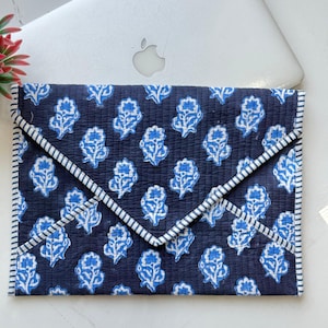 May include: A quilted envelope-style laptop sleeve featuring a dark navy blue fabric with a repeating light blue floral block print pattern. The edges are finished with a white and navy blue striped fabric trim, creating a distinct border.