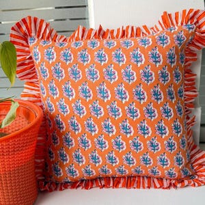 May include: Square orange pillow with a repeating pattern of blue and pink floral designs. The pillow has a ruffled edge in orange and white stripes. The pillow is a decorative home accent.