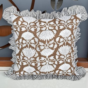 May include: Square decorative pillow with a brown floral pattern and white accents. The pillow has a ruffled border with black and white stripes. The pillow is designed for home decor.