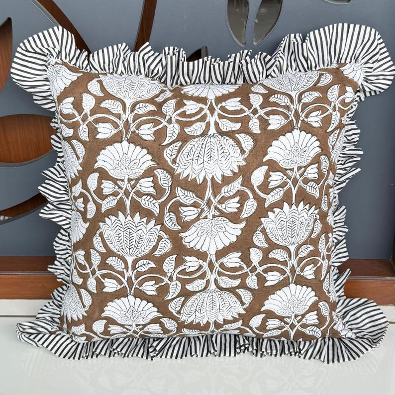 May include: Square decorative pillow with a brown floral pattern and white accents. The pillow has a ruffled border with black and white stripes. The pillow is designed for home decor.