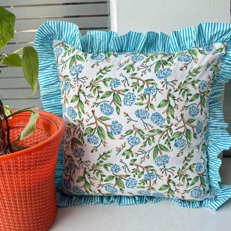 May include: Square decorative pillow with a white background and a floral pattern in blue and green. The pillow has a ruffled border in a blue and white striped fabric. An orange pot with a plant is in the foreground.