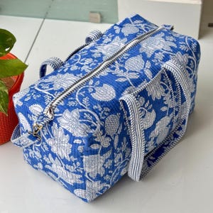 Può includere: Una borsa da viaggio blu e bianca con motivo floreale e cerniera argentata. La borsa ha due manici e una tracolla staccabile. Il motivo floreale è bianco su sfondo blu.