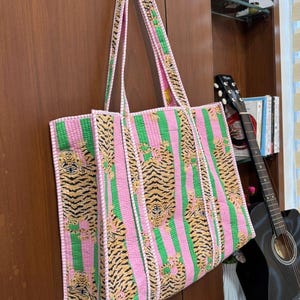May include: A large tote bag with a pink and green striped pattern, featuring tiger print accents. The bag has long handles and is made of a quilted fabric. The bag is hanging on a wooden door.