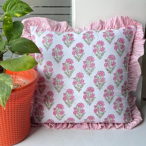 May include: Square decorative pillow with a white background and a repeating pattern of pink floral designs. The pillow has a ruffled edge with pink and white stripes. A potted plant is visible in the background.