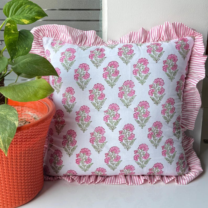 May include: Square decorative pillow with a white background and a repeating pattern of pink floral designs. The pillow has a ruffled edge with pink and white stripes. A potted plant is visible in the background.