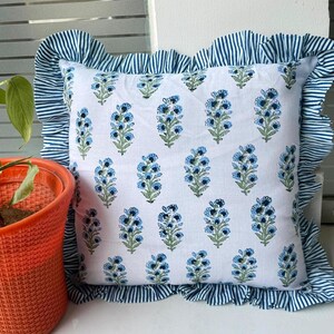 May include: Square white pillow with a blue floral pattern and a ruffled border with blue and white stripes. The pillow is a decorative item, likely for a bed or sofa.