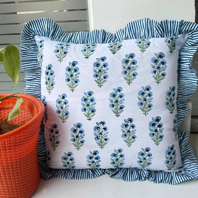 May include: Square white pillow with a blue floral pattern and a ruffled border with blue and white stripes. The pillow is a decorative item, likely for a bed or sofa.