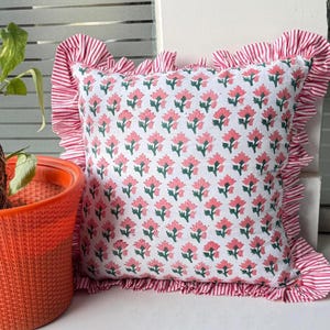 May include: Square white pillow with a pink floral pattern and green leaves. The pillow has a ruffled edge with red and white stripes. The pillow is placed near an orange pot with a green plant.