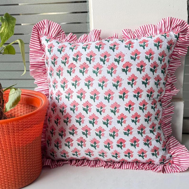 May include: Square white pillow with a pink floral pattern and green leaves. The pillow has a ruffled edge with red and white stripes. The pillow is placed near an orange pot with a green plant.