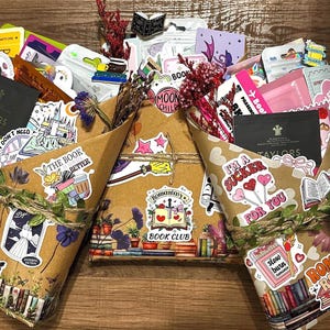 Pu&ograve; includere: Tre pacchi di libri avvolti in carta marrone, decorati con vari adesivi colorati con testi come "Romantasy Book Club", "I'm a sucker for you" e "Romance Reader", arricchiti da fiori secchi e legati con spago rustico.