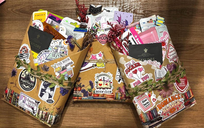 Pu&ograve; includere: Tre pacchi di libri avvolti in carta marrone, decorati con vari adesivi colorati con testi come "Romantasy Book Club", "I'm a sucker for you" e "Romance Reader", arricchiti da fiori secchi e legati con spago rustico.