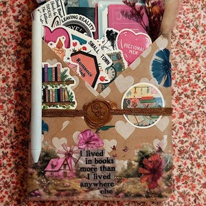 Pu&ograve; includere: Una busta di carta marrone decorata con cuori bianchi, adesivi e un sigillo di ceralacca raffigurante una rosa. La busta contiene vari articoli di cancelleria, tra cui adesivi e fiori secchi. Uno stilo bianco &egrave; fissato sul lato. Il testo recita: I lived in books more than I lived anywhere else.