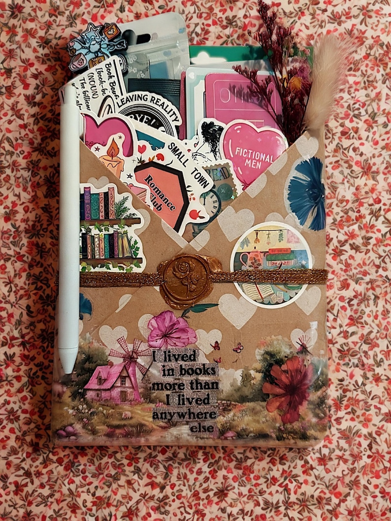 Pu&ograve; includere: Una busta di carta marrone decorata con cuori bianchi, adesivi e un sigillo di ceralacca raffigurante una rosa. La busta contiene vari articoli di cancelleria, tra cui adesivi e fiori secchi. Uno stilo bianco &egrave; fissato sul lato. Il testo recita: I lived in books more than I lived anywhere else.