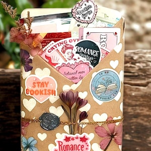 Pu&ograve; includere: Una busta rettangolare in carta kraft decorata con cuori bianchi e accenti floreali, contenente vari adesivi e cartoline a tema librario. Il testo visibile include "Stay Bookish", "Romance reader", "Crying over fictional men" e "In my bookish era".
