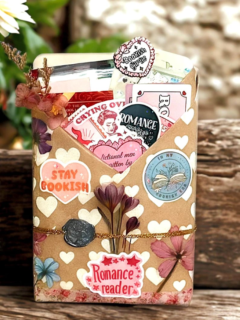 Pu&ograve; includere: Una busta rettangolare in carta kraft decorata con cuori bianchi e accenti floreali, contenente vari adesivi e cartoline a tema librario. Il testo visibile include "Stay Bookish", "Romance reader", "Crying over fictional men" e "In my bookish era".