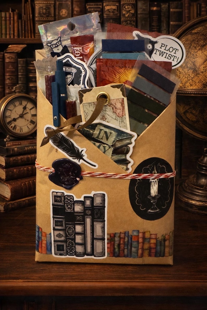 Pu&ograve; includere: Una busta di carta marrone riempita di articoli di cancelleria, tra cui una penna blu, adesivi di libri, una piuma e una candela. La busta &egrave; legata con uno spago rosso e bianco e presenta un sigillo di ceralacca, un'etichetta mappa e PLOT TWIST.