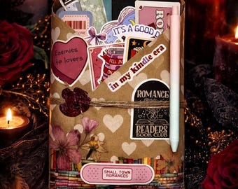 Può includere: Un cofanetto regalo avvolto in carta marrone decorato con adesivi e una penna. Gli adesivi includono le frasi "Enemies to lovers" e "Small Town Romances". La scatola è legata con spago e circondata da candele e rose.
