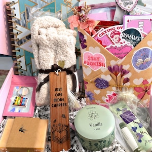 Pu&ograve; includere: Una confezione regalo contenente un taccuino a spirale con motivo geometrico verde acqua, un paio di calzini beige soffici, un segnalibro in legno con la scritta "JUST ONE MORE CHAPTER", una candela alla vaniglia, una saponetta, un portachiavi e una busta decorata.