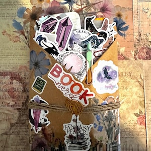 Pu&ograve; includere: Una busta di carta marrone riempita con adesivi assortiti raffiguranti cristalli, draghi e motivi celesti. La busta &egrave; legata con dello spago e riporta scritte tra cui "BOOK nerd", "you are magic" e "FANTASY READER" con una piccola clip verde.
