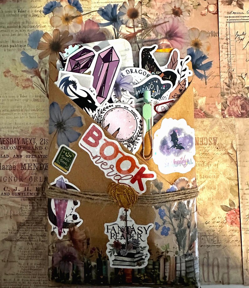 Pu&ograve; includere: Una busta di carta marrone riempita con adesivi assortiti raffiguranti cristalli, draghi e motivi celesti. La busta &egrave; legata con dello spago e riporta scritte tra cui "BOOK nerd", "you are magic" e "FANTASY READER" con una piccola clip verde.