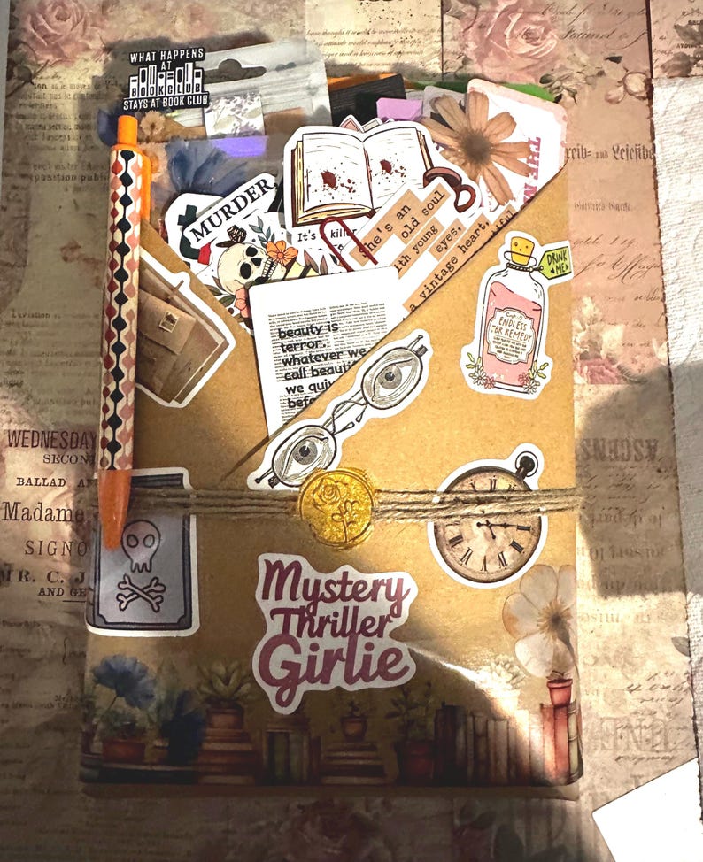 Pu&ograve; includere: Una busta di carta marrone riempita con vari adesivi tra cui un teschio, un libro aperto, occhiali, un orologio e una boccetta. La busta &egrave; legata con dello spago, presenta un sigillo di ceralacca dorato e il testo Mystery Thriller Girlie.