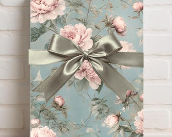 Puede incluir: Una caja de regalo rectangular envuelta en papel floral con peonías rosas y follaje verde sobre un fondo azul claro. Una cinta de raso plateada está atada en un lazo alrededor de la caja. La caja está colocada sobre una superficie de madera.