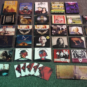 May include: A selection of music CDs, displaying album covers with varied artwork and band names. The CDs are arranged on a dark surface, showcasing a range of genres and artists. The image includes albums by Metallica, Moby, and other bands.