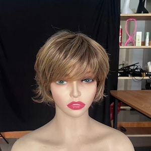 Könnte beinhalten: Eine kurze, geschichtete Perücke mit blonden und braunen Strähnen. Die Perücke ist mit einem Seitenscheitel und einem Pony gestylt. Die Perücke befindet sich auf einem Mannequin-Kopf. Der Text "BAG wig" ist auf die Brust des Mannequins geschrieben.