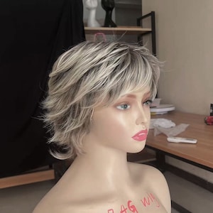 Könnte beinhalten: Eine kurze, zerzauste blonde Perücke mit hellbraunen Strähnen. Die Perücke ist mit einem Seitenscheitel gestylt und hat einen geschichteten, strukturierten Look. Die Perücke befindet sich auf einem Mannequin-Kopf mit dem Text "BAG WIG" auf der Brust.