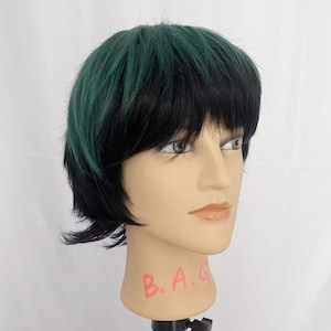 May include: A mannequin head with a short, layered wig. The wig features a combination of black and dark green hair. The mannequin is mounted on a black stand against a white background. The letters "B.A.G" are written in red on the mannequin's neck.