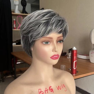 Può includere: Una parrucca corta, grigia e nera su una testa di manichino. La parrucca ha uno stile a strati e irregolare. Il manichino indossa un rossetto rosso. Il testo "BAG wig" è scritto sul petto del manichino.