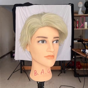Puede incluir: Una cabeza de maniquí con cabello corto rubio claro. El maniquí lleva una peluca y las letras "B.A.G." escritas en la parte delantera de la cabeza.
