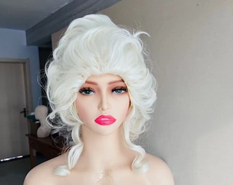May include: A white wig with a large, bouffant style. The wig is styled with loose curls and is worn on a mannequin head. The mannequin is wearing a pink shirt and has a label that says "BAG COS" on the chest.