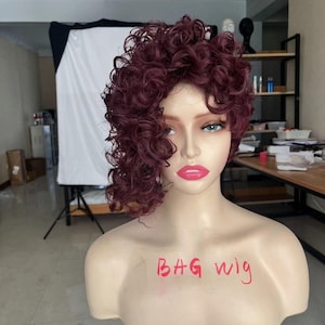 Op de afbeelding: Een bordeauxrode krullende pruik op een mannequinhoofd. De pruik is gestyled met een scheiding aan de zijkant en heeft veel volume. De mannequin draagt roze lippenstift en heeft groene ogen. De tekst "BAG wig" is op de borst van de mannequin geschreven.