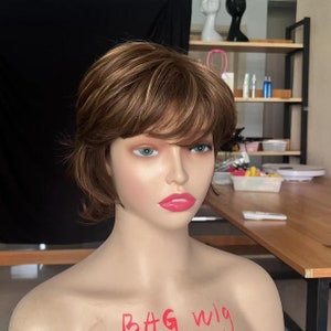 Puede incluir: Una peluca corta marrón con reflejos rubios en una cabeza de maniquí. La peluca tiene raya lateral y flequillo. El maniquí lleva un pintalabios rosa. El texto "Bag wig" está escrito en el pecho del maniquí.