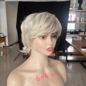 May include: A short, platinum blonde wig with a side part and choppy layers. The wig is styled on a mannequin head with the text "BAG wig" written on the chest.