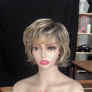 Puede incluir: Una peluca rubia corta y con capas. La peluca tiene una base marrón claro con reflejos rubios. La peluca está peinada con raya lateral y flequillo. La peluca está en una cabeza de maniquí. El texto "BAG wig" está escrito en el pecho del maniquí.