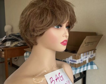 May include: A mannequin head displaying a short, brown wig with a layered style and side-swept fringe. The wig is styled with soft waves. The mannequin has painted features, including blue eyes and red lipstick. A handwritten note on the mannequin reads "BAG wigs".