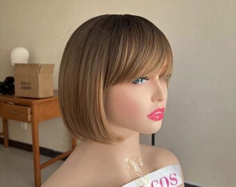 May include: A brown and blonde bob wig with bangs. The wig is on a mannequin head. The mannequin is wearing a white shirt with a pink sticker that says "BAG COS" on it.
