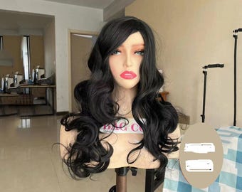 May include: A black, wavy wig styled on a mannequin head. The wig features long, flowing curls and a natural-looking hairline. The mannequin has a fair complexion, red lips, and is mounted on a stand. The background is a room with a table and equipment.