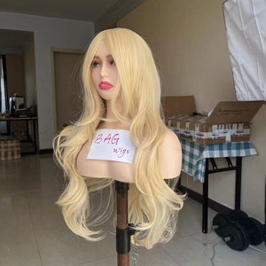 Puede incluir: Una peluca larga, ondulada y rubia en una cabeza de maniquí. La peluca está peinada en ondas sueltas y tiene un color de aspecto natural. El maniquí lleva una camisa blanca con las palabras "BAG wigs" escritas en ella.