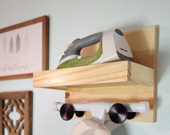 May include: A white and gray ironing board cover hangs on a wooden shelf with a white iron on top. The shelf is mounted to a light blue wall.