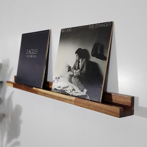 May include: A wooden record shelf with two vinyl record albums displayed. One album is black with the text "EAGLES THE LONG RUN". The other album is black and white with the text "BILLY JOEL THE STRANGER". The shelf is mounted on a white wall.