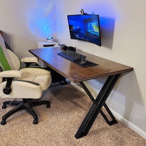 May include: A modern desk with a dark wood top and black X-shaped legs. A green and white gaming chair sits in front of the desk. A curved monitor with a blue light is mounted to the wall above the desk.