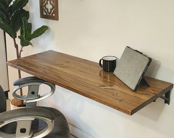 May include: A brown wooden wall-mounted desk with a black mug and a tablet on top. The desk is mounted to a white wall with black metal brackets. A gray office chair is in the foreground.