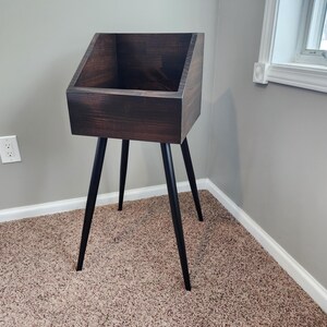 May include: A dark stained wooden record stand with a rectangular top and angled sides. The stand is supported by three black, tapered legs. The stand is placed in a corner of a room with a neutral color scheme.