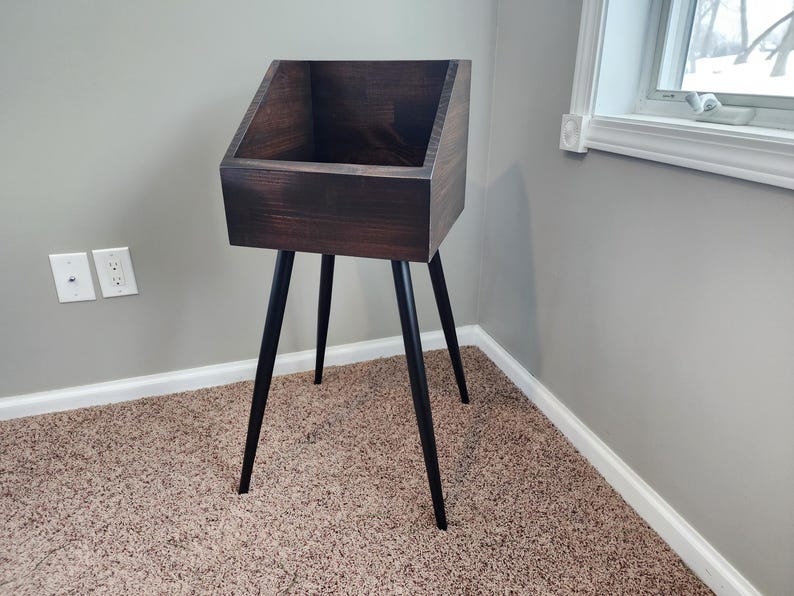 May include: A dark stained wooden record stand with a rectangular top and angled sides. The stand is supported by three black, tapered legs. The stand is placed in a corner of a room with a neutral color scheme.
