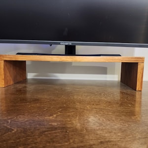 May include: A wooden monitor stand with a natural wood finish. The stand is made of two pieces of wood that are joined together to form a rectangular shape. The stand is sitting on a brown surface.
