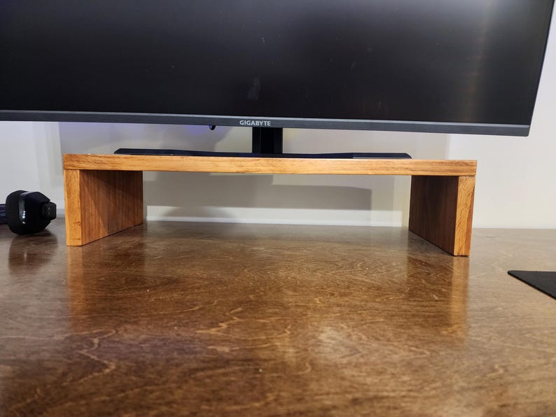 May include: A wooden monitor stand with a natural wood finish. The stand is made of two pieces of wood that are joined together to form a rectangular shape. The stand is sitting on a brown surface.